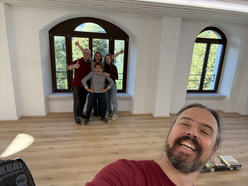 Eine fröhliche Gruppe von fünf Personen steht in einem neu eingerichteten Raum mit großen Fenstern und Holzfußboden. Der Raum ist hell und bietet einen Blick ins Freie, während eine Person im Vordergrund ein Selfie macht.