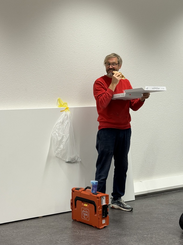 Ein Mann in einem roten Pullover hält eine Pizza und steht neben einem weissen Wandpaneel in einem neu eingerichteten Atelier. Auf dem Boden stehen Werkzeuge und Materialien, während im Hintergrund eine Tüte hängt.